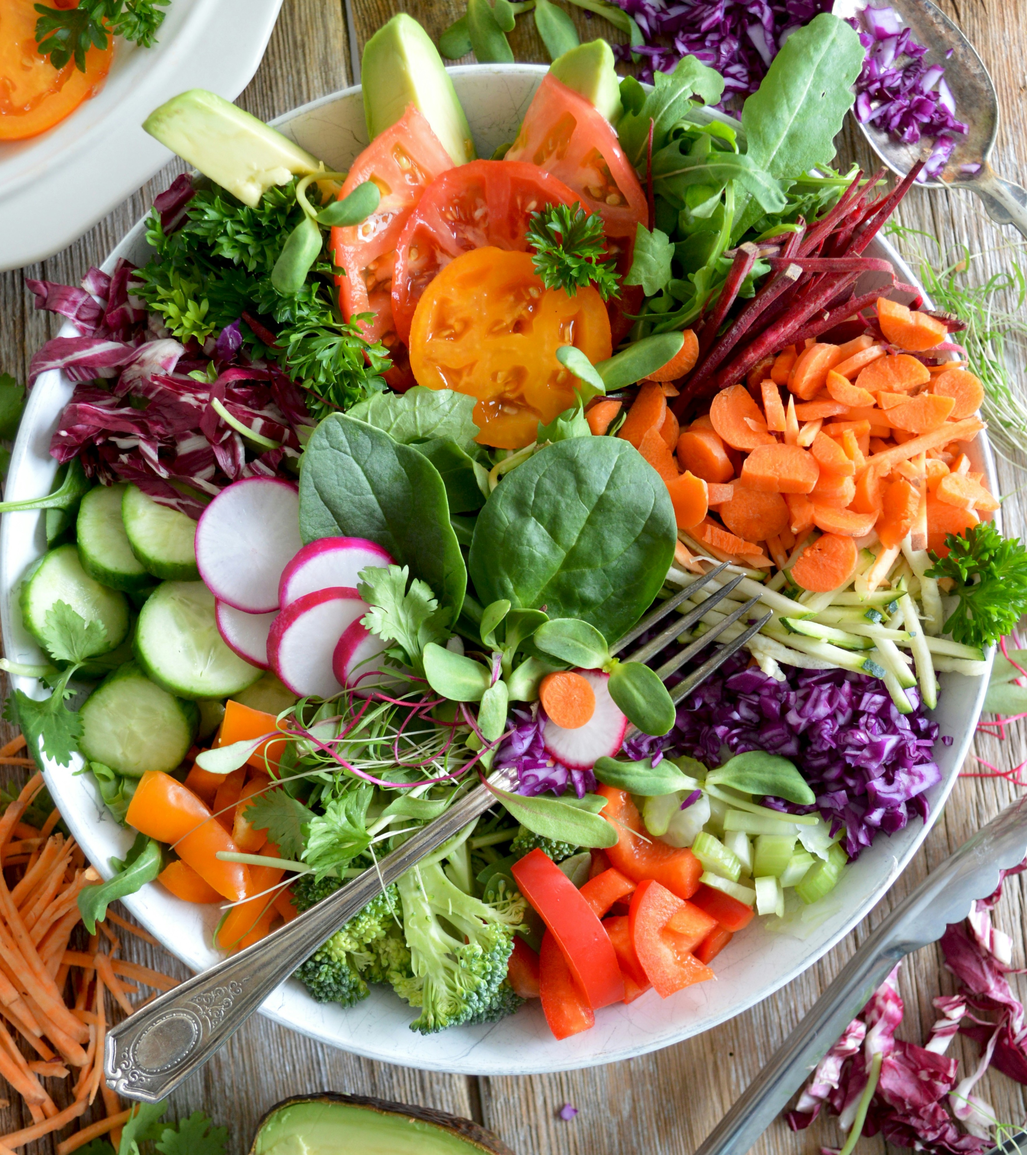 Top-down view of a white bowl packed with colourful raw salad vegetables including spinach, avocado, carrots, beets, and cabbage on a wooden table